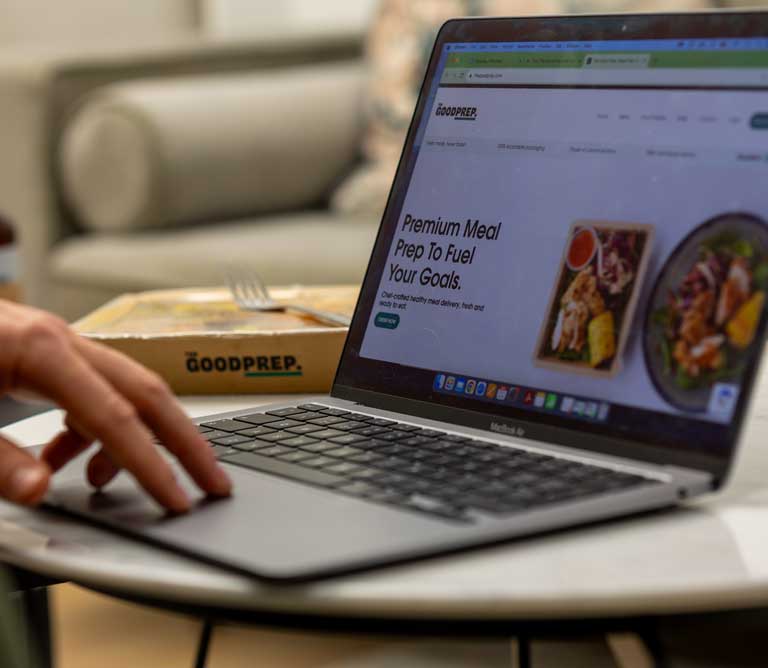 The image shows a person sat at a laptop, looking at a premium meal delivery service website.