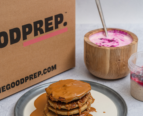 Brown recyclable box with 'The Good Prep' logo, next to a plate of stacked pancakes and a bowl of overnight oats.